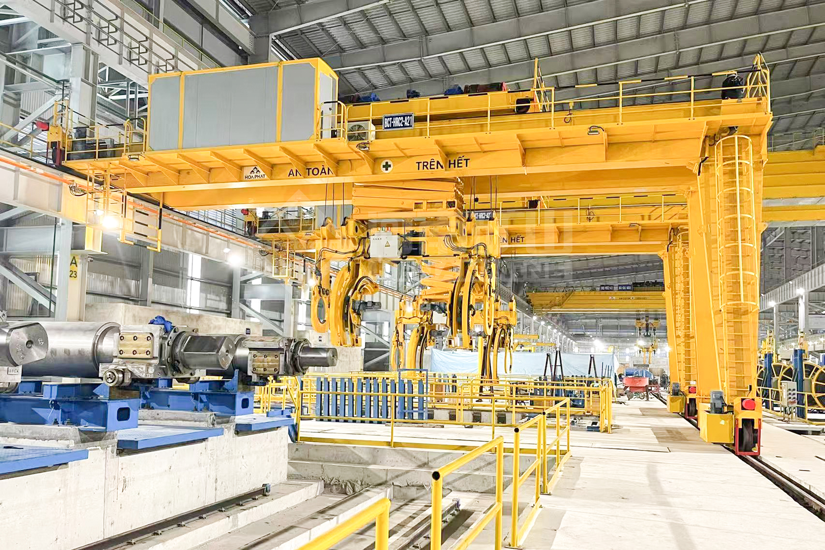 Yellow overhead crane with specialized lifting attachment above steel-processing equipment
